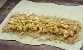 Слоеное тесто, заранее размороженное, разложите на бумаге для выпечки или на разделочной доске и намного раскатайте. Листы теста равномерно посыпьте смесью орехов с сухарями. Поверх разложите подготовленную яблочную начинку.