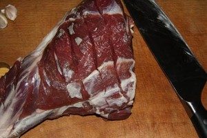 Кусок мяса баранины хорошо промойте под проточной водой и обсушите полотенцем. Затем сделайте на мясе несколько глубоких надрезов. Почистите чеснок и порежьте его продольными кусочками. Поместите чеснок в надрезы на мясе.
