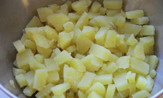 Картофель промойте и отварите «в мундирах» до готовности. Затем его остудите, очистите от кожуры и нарежьте небольшими кубиками. Нарезанный картофель сразу переложите в салатницу.