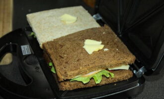 Выкладываем сэндвичи в мультипекарь, а поверх каждого бутерброда кладем по небольшому кусочку масла для сливочного вкуса.