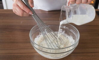 Теперь готовим тесто для блинов. В большую ёмкость просеиваем муку с содой и солью через сито. Затем добавляем туда сахарный песок и всё перемешиваем. Далее берём венчик и, не переставая помешивать, вливаем молоко и 250 мл тёплой воды.