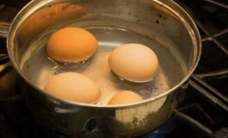 Выкладываем яйца (заранее достаем из холодильника, чтобы они стали комнатной температуры) в кастрюлю и заливаем теплой водой. Слегка подсаливаем воду. Все эти манипуляции необходимы для того, чтобы яйца не трескались в процессе варки. Когда вода закипит, варим яйца около десяти минут до готовности. Затем сливаем кипяток и остужаем яйца в холодной воде.