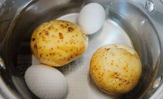 Одновременно отварить промытый в мундирах картофель. Куриные яйца отварить вкрутую. Отваренные ингредиенты быстро остудить в холодной воде.