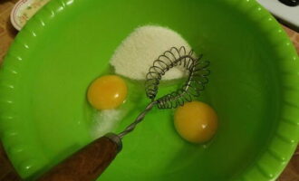 В посуду для замеса теста разбейте два куриных яйца. Насыпьте к ним, согласно рецепту, сахар с солью и венчиком взбейте в пышную массу.