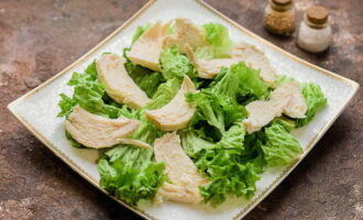 Подготовьте блюдо, на котором будете собирать салат. Листья салата сполосните и просушите. Застелите блюдо, а сверху распределите нарезанные куриные ломтики.