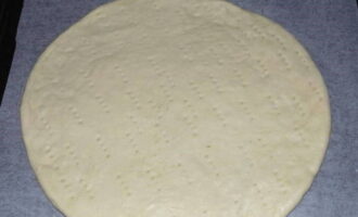 Тесто для пиццы тонко раскатываем в круг и выкладываем на противень с пергаментом. По всей поверхности делаем проколы вилкой или зубочисткой.