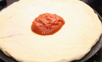 После повторного подъема тесто еще раз обомните, раскатайте в тонкую лепешку толщиной не более 1 см, соответственно размеру выбранной формы для выпечки. Тесто равномерно покройте тонким слоем соуса для пиццы.