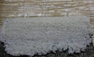 Для сборки ролла на бамбуковый коврик положите лист нори. Поверх его ровным слоем уложите сваренный рис и ковриком его хорошо уплотните.