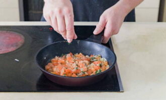 Отправляем к луку кусочки помидоров и измельченный базилик. Солим содержимое и посыпаем черным молотым перцем. Томим овощи до испарения жидкости.