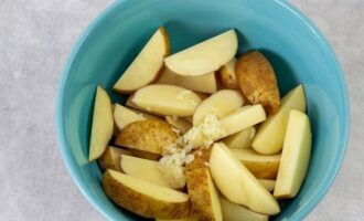 Картофель не очищайте. Нарежьте клубни тонкими дольками и переложите в объемную миску. Чесночные зубчики очистите от шелухи, затем пропустите через пресс и полученную кашицу добавьте в миску к картофелю.