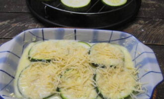 На дно формы для выпекания вливаем немного соуса. Выкладываем часть кругов кабачков, снова поливаем соусом и посыпаем тертым сыром.