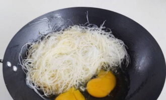 С рисовой лапши слейте воду. Затем выложите ее в вок и разбейте куриные яйца. Когда яйца немного схватятся, перемешайте их с лапшой.