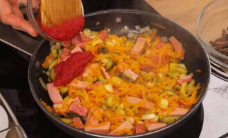 Глубокую сковороду устанавливаем на конфорку. Разогреваем. Наливаем растительное масло, выкладываем лук и морковку. Нарезаем огурцы и добавляем к овощам. Кладем оба вида колбасы и мясо. Перемешав, тушим при постоянном помешивании 6 минут. После чего приправляем томатной пастой. Перемешиваем до однородности и готовим еще пару минут.